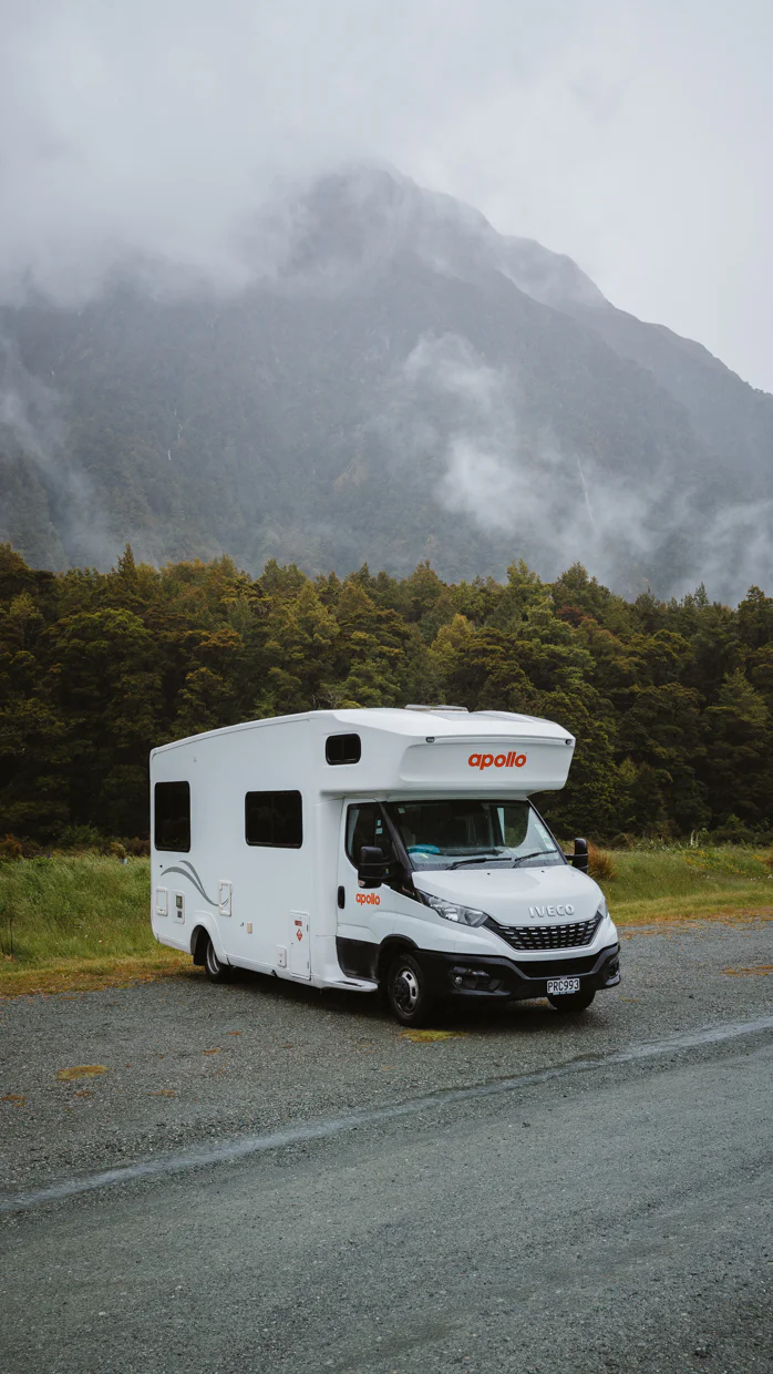 White recreational vehicle parked on a gravel road with forest and misty mountains in the background.