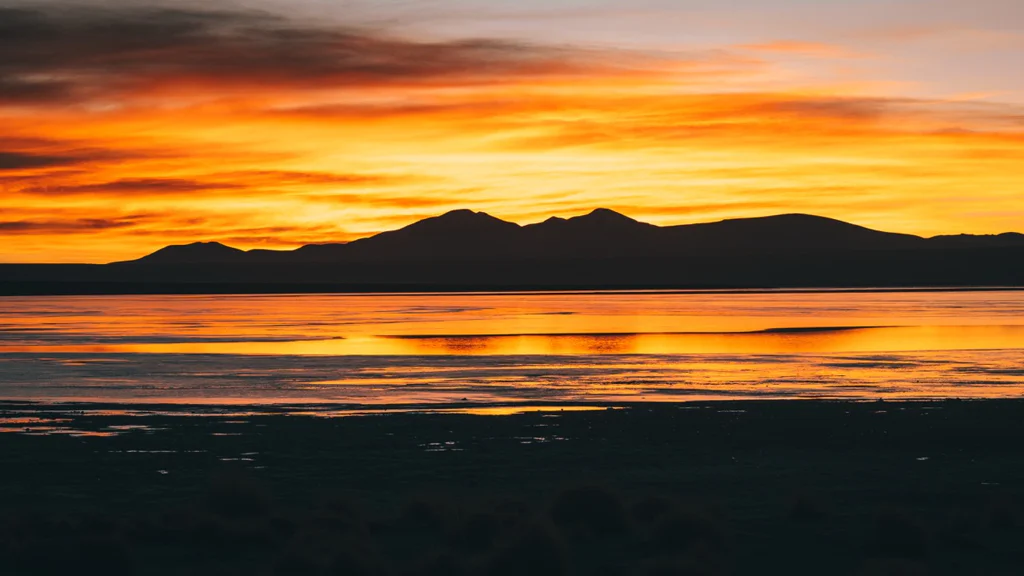 A vibrant orange and yellow sunset over a lake with dark mountain silhouettes.