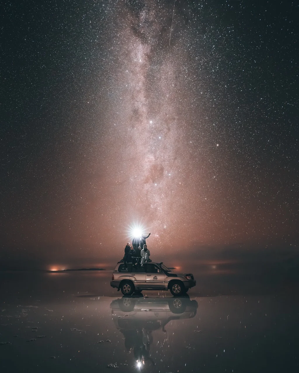 Group of people on a car under a starry night sky with the Milky Way reflected on a calm surface.