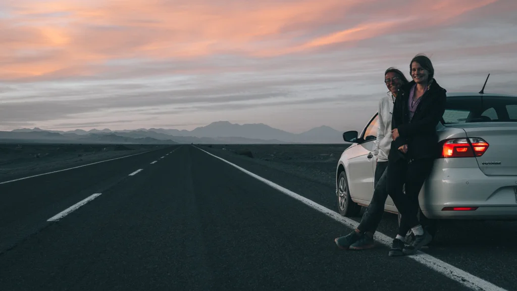 Two people leaning against a white car on the side of an empty road at sunset with mountains in the background.