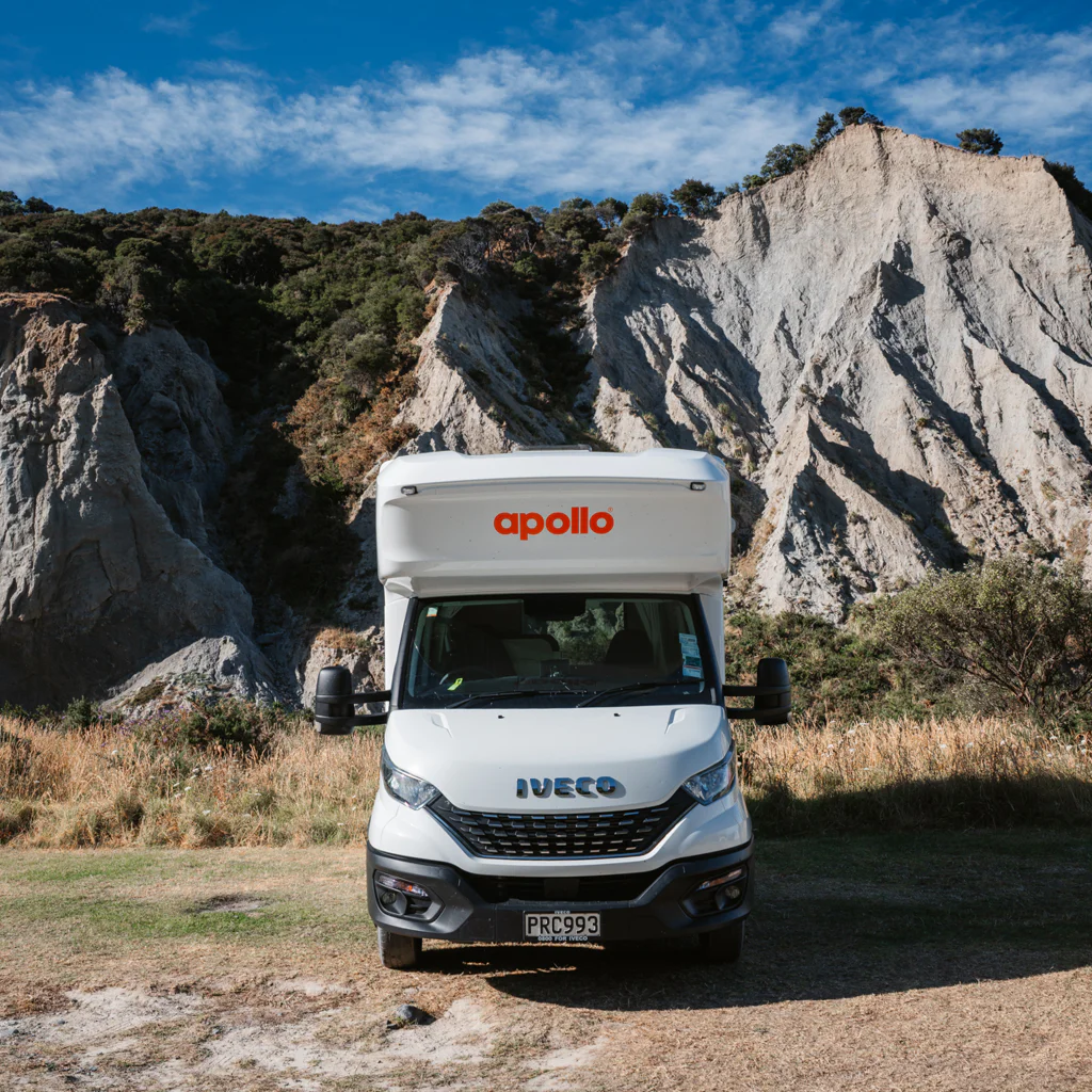 Front view of a white recreational vehicle parked in a natural mountainous area under a blue sky.