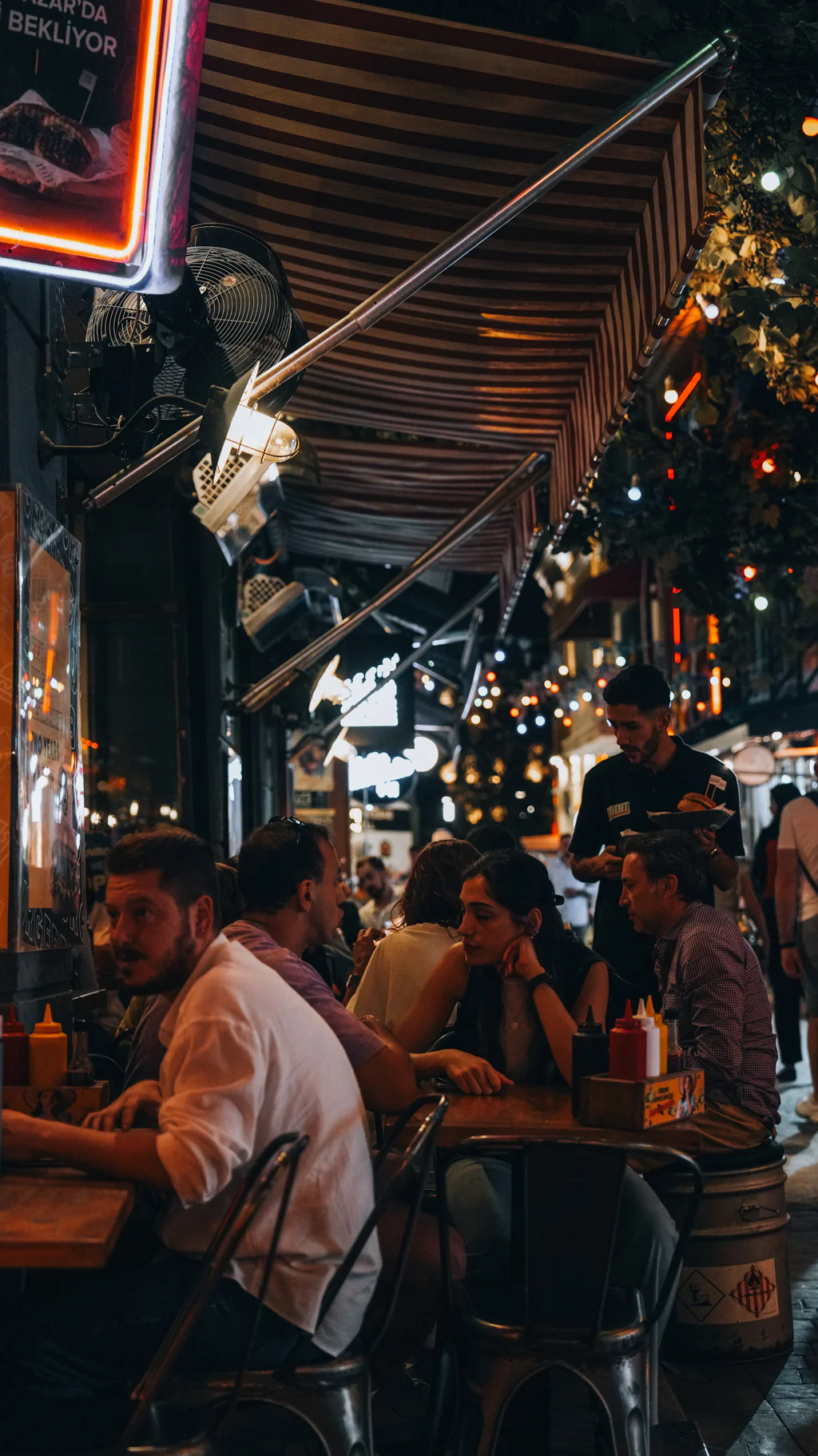 People sitting and talking at an outdoor restaurant under striped awnings and warm string lights at night.