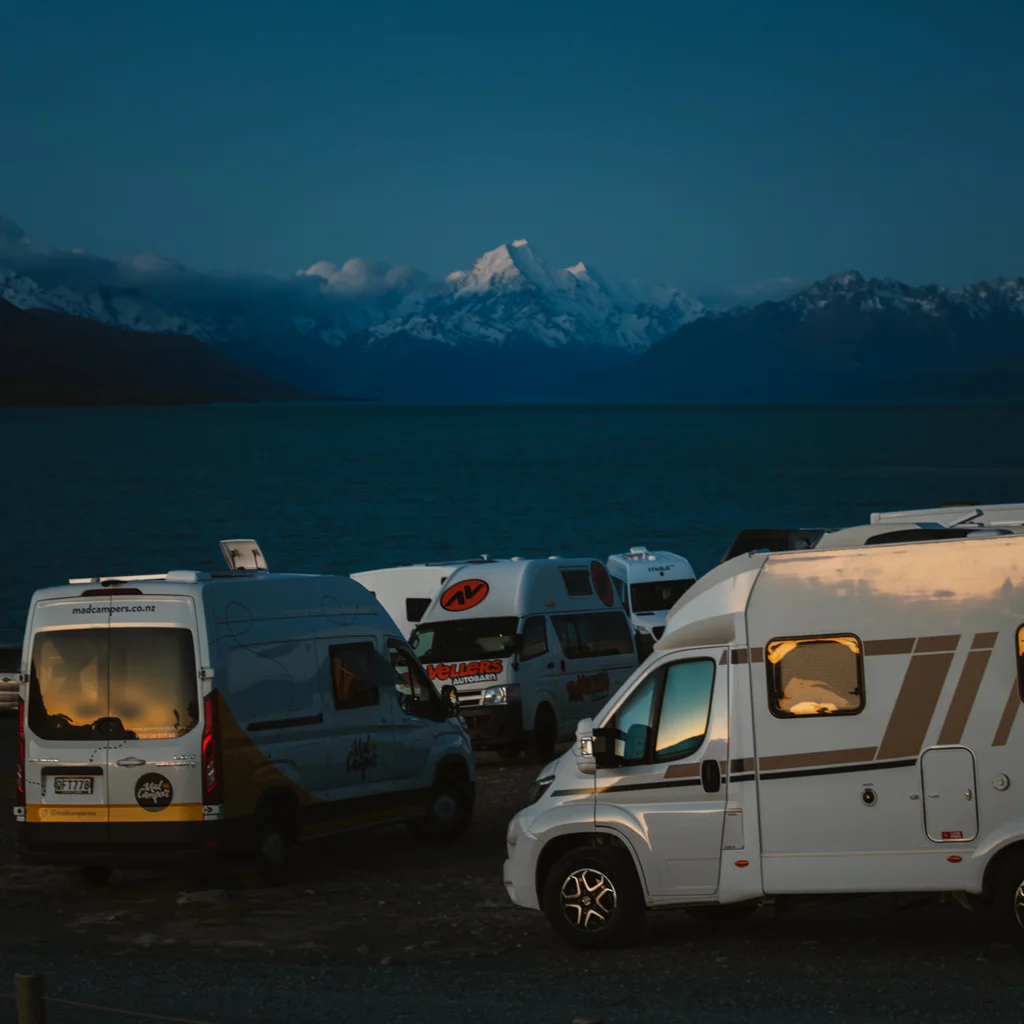 Camping vans parked by a lake with snow-covered mountains in the background at dusk.