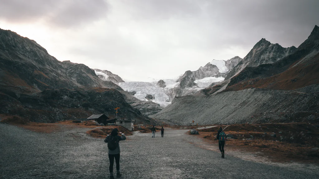Hikers walking on a gravel path toward snowy mountains under an overcast sky.