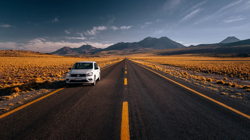 White car parked on the side of a long road in a dry mountainous landscape under a blue sky.