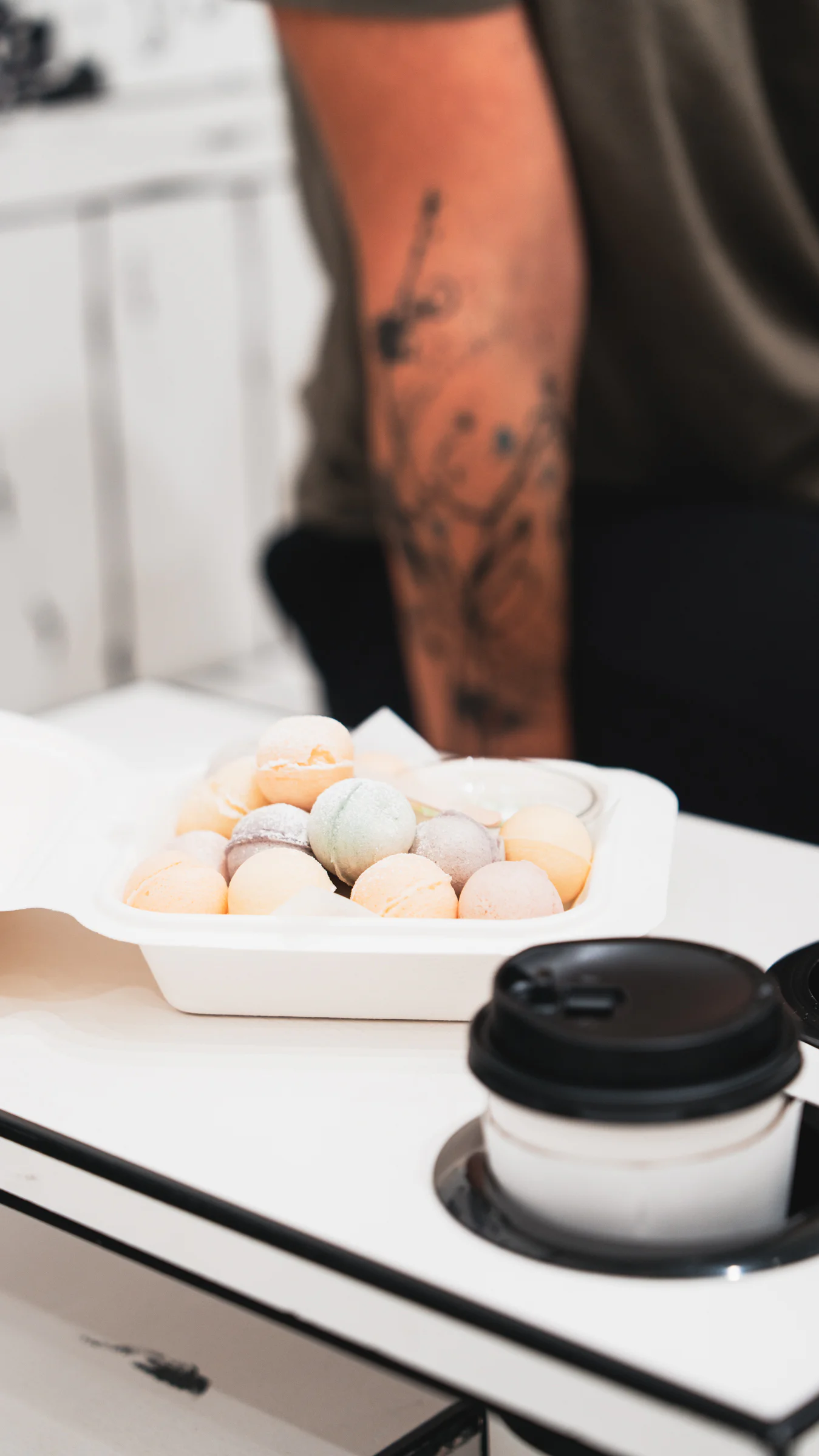 Close-up of pastel macarons in a white tray next to a coffee cup with a tattooed arm blurred in the background.