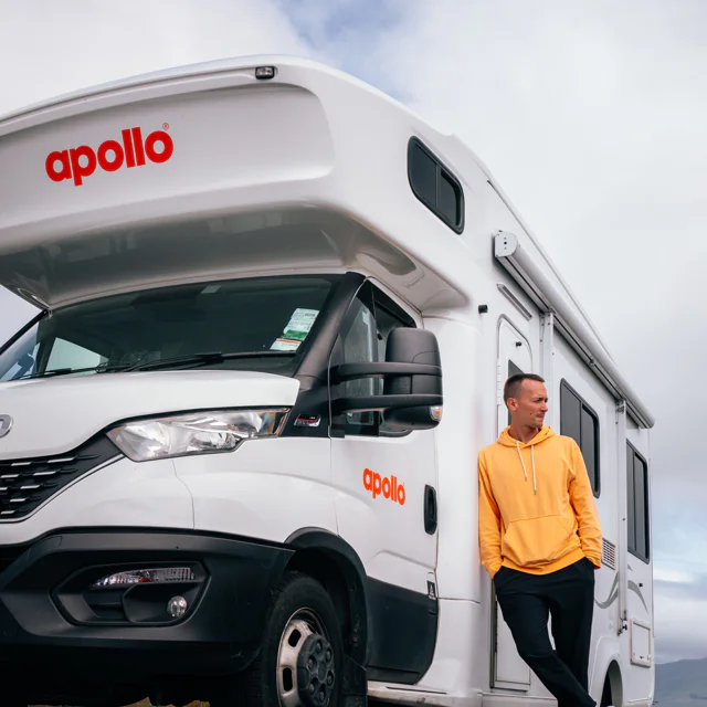 Man in yellow hoodie leaning against a white Apollo camper van under a cloudy sky.