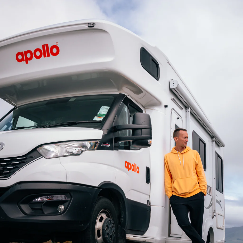 Man in yellow hoodie leaning against a white Apollo camper van under a cloudy sky.