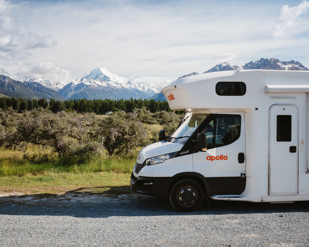 White recreational vehicle parked by a gravel road with mountains and trees in the background.