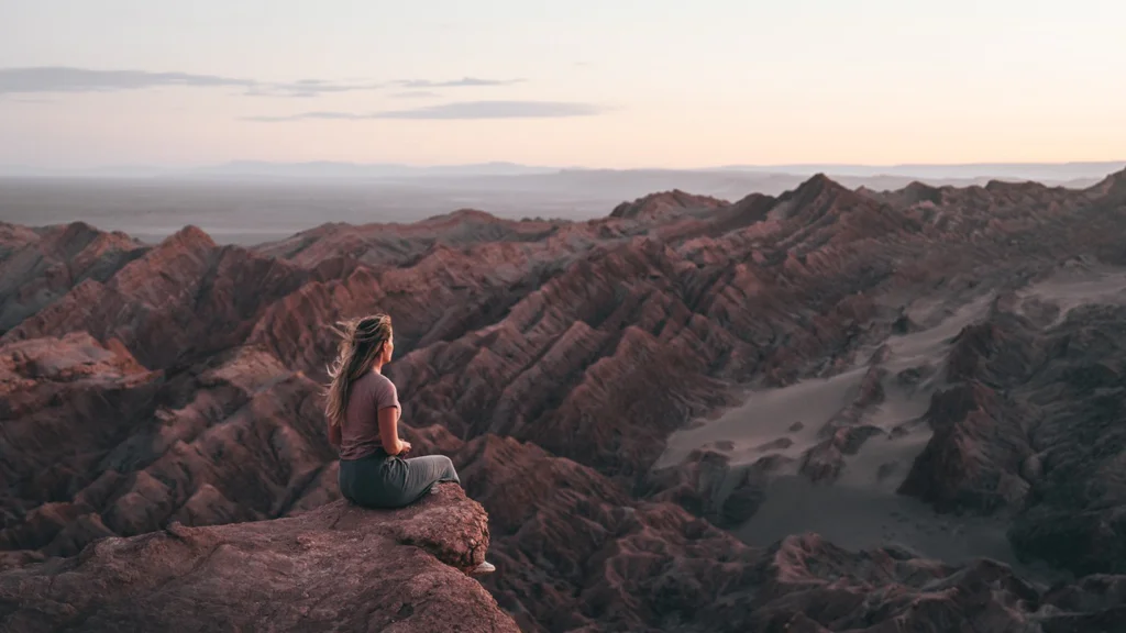 Woman sitting on a rock overlooking a mountainous desert landscape at sunset.