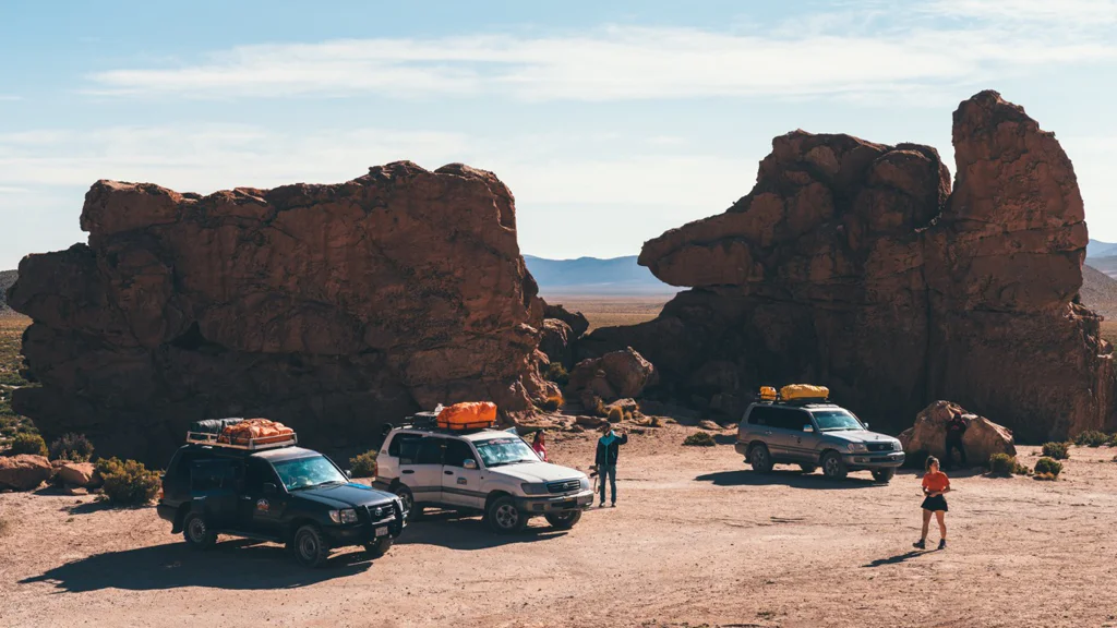 Three off-road vehicles and two people in a desert landscape with large rock formations.