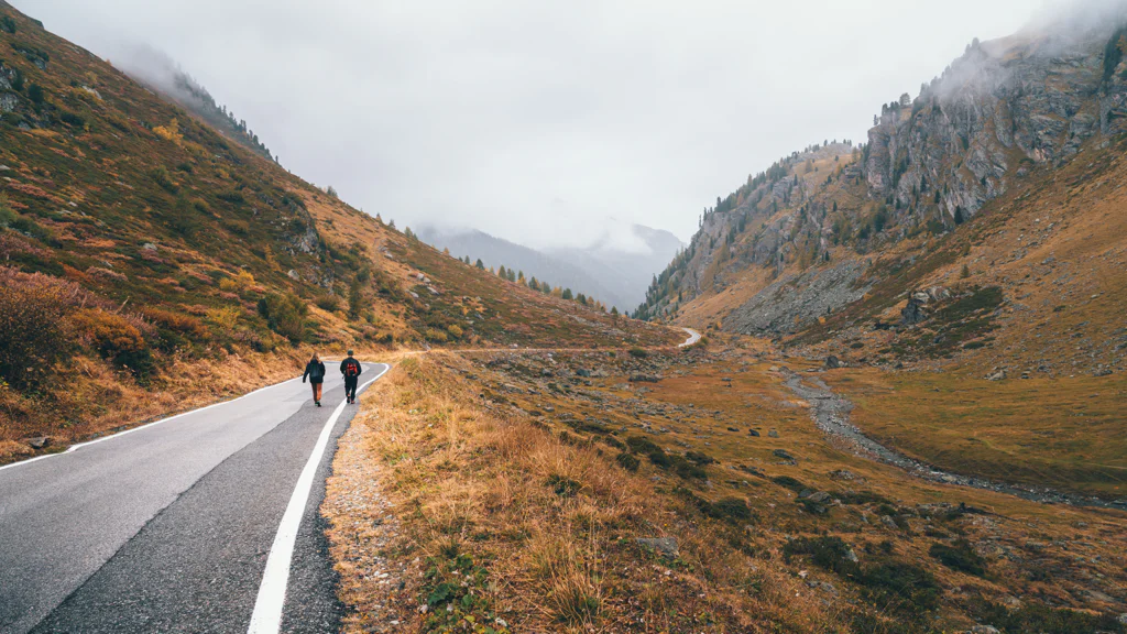 Two people walking on a winding road through a mountain valley with autumn foliage.