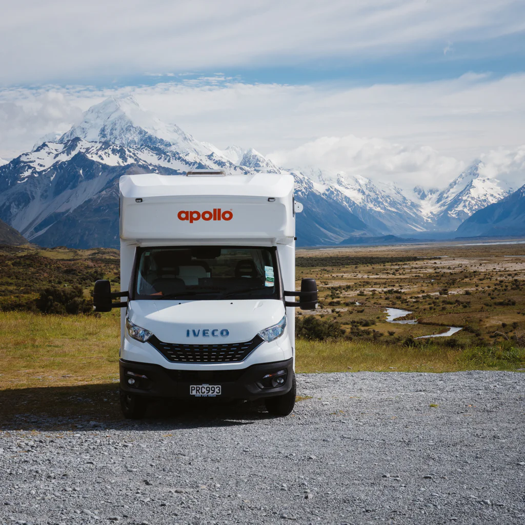 White recreational vehicle parked in front of snow-covered mountains under a blue sky.