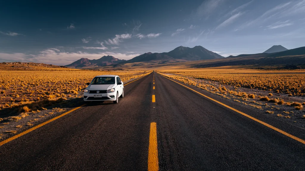 White car parked on a long road through a dry landscape with mountains in the background.