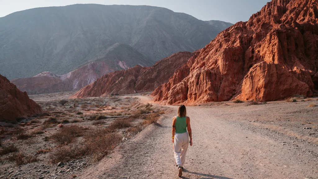 Person walking on a dirt path through red rocky mountains in a desert landscape.