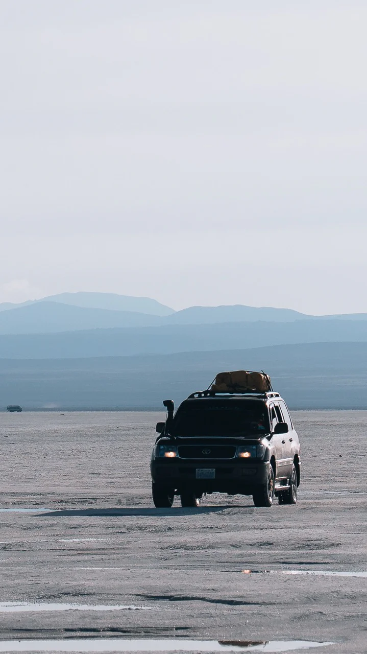 Black SUV driving on a flat desert landscape with mountains in the background under an overcast sky.