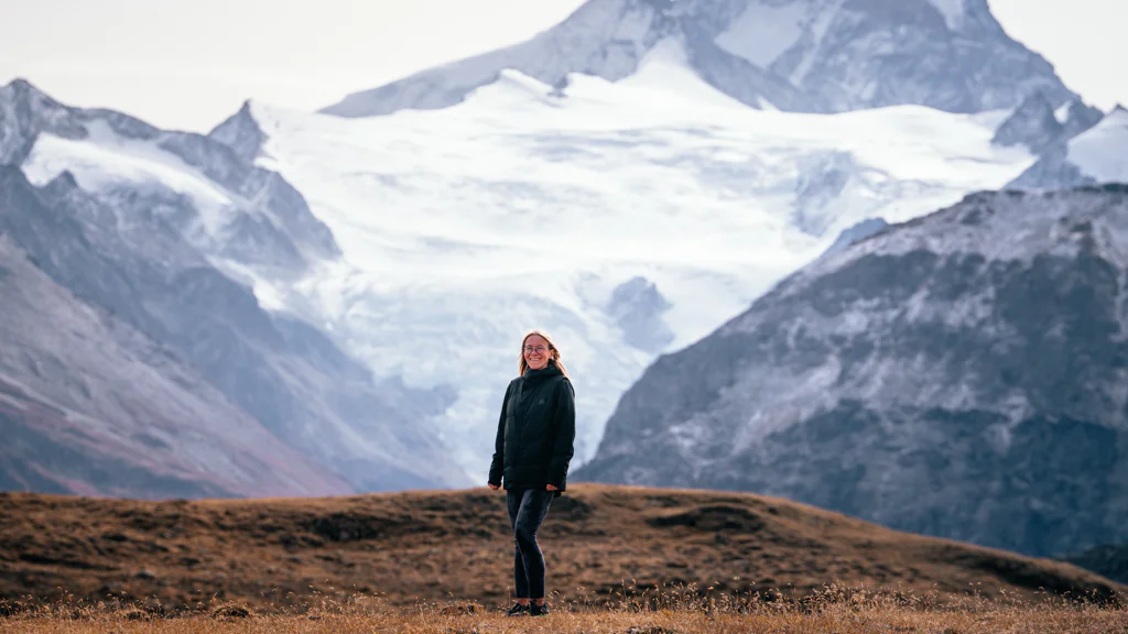 Person standing on a grassy hill with snow-covered mountains in the background.