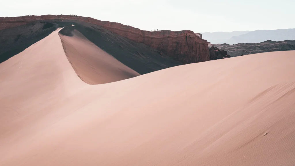 Wide view of smooth sand dunes with rocky cliffs in the background under a pale sky.