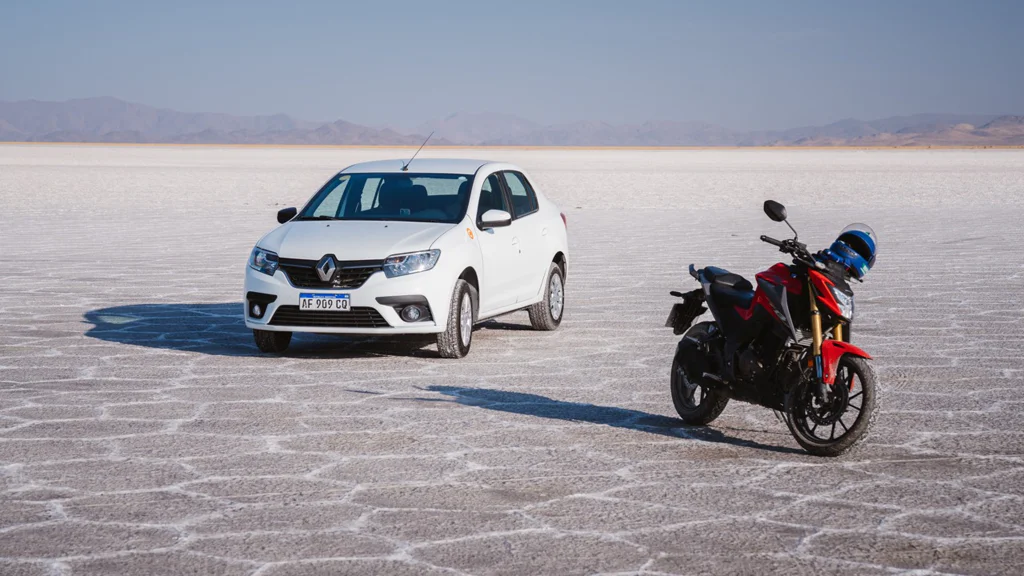 A white Renault car and a red and black motorcycle parked on a salt flat with mountains in the distance.