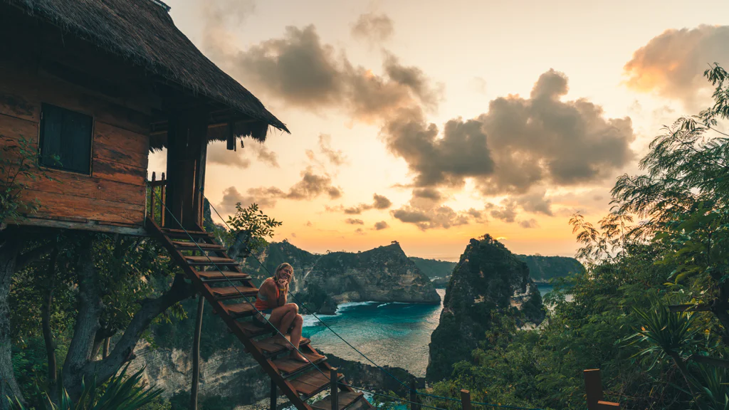 Person sitting on stairs of a treehouse overlooking a coastal sunset landscape with rock formations and greenery.