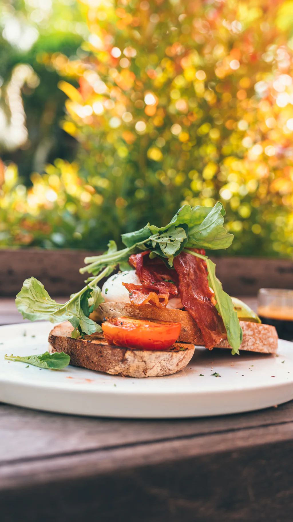 Close-up of a breakfast plate with toast, bacon, poached egg, tomato, and greens on a white plate outdoors.