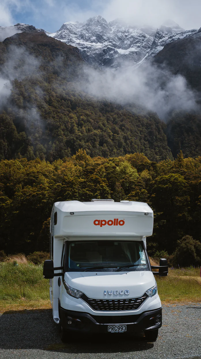 White Apollo recreational vehicle parked in front of a forest with misty mountains in the background.