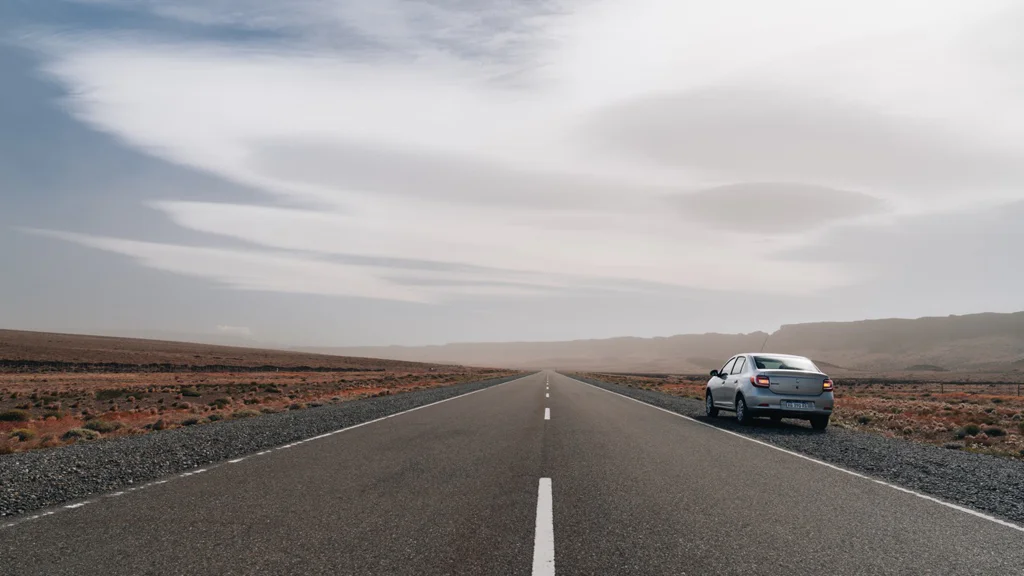 A silver car parked beside a long, empty road in a desert landscape under a cloudy sky.