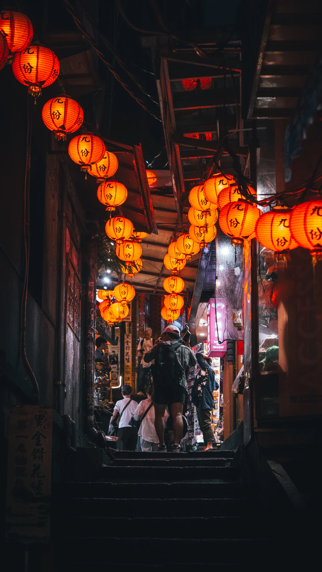 People walking up stairs under glowing orange lanterns in a narrow Jioufen alley at night.