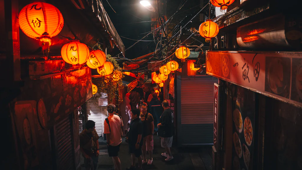 Nighttime street in Jioufen lit by orange lanterns with people walking through a narrow alley.